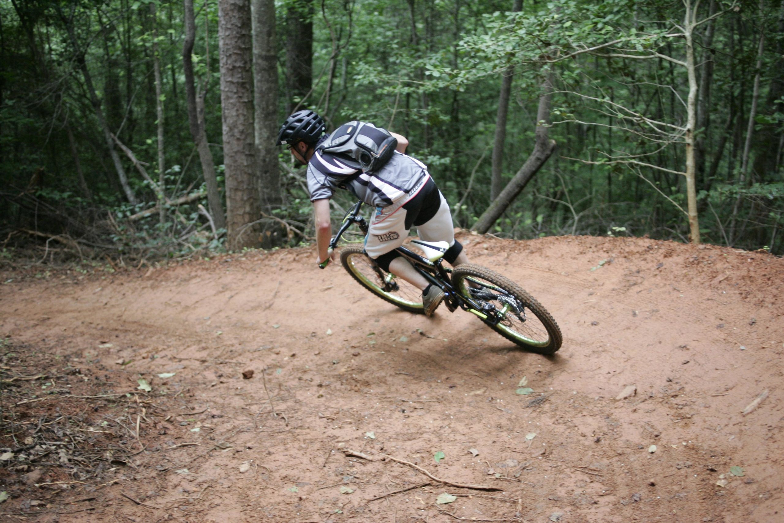 A person riding a mountain bike on a dirt trail in a forested area, leaning into a curve. The cyclist is wearing a helmet and biking gear, with trees and foliage visible in the background. Dark Mountain Trail mountain bike trail.