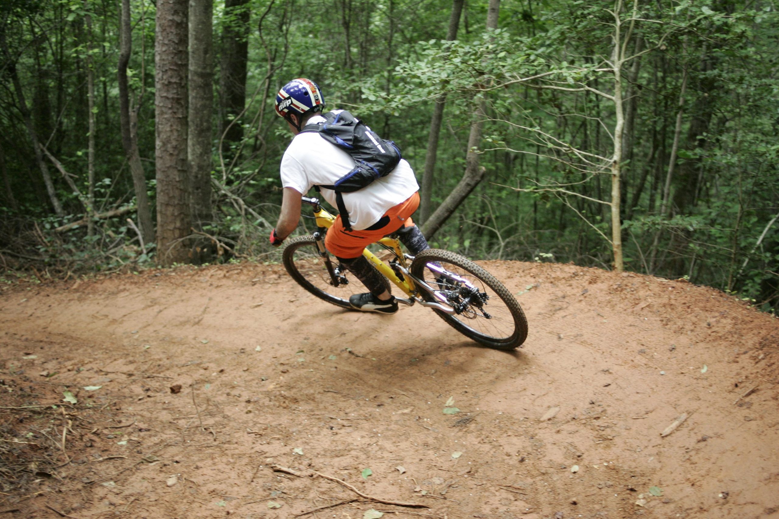 A mountain biker in a white shirt and orange shorts navigates a curved dirt trail through a dense forest. The biker, wearing a helmet and a backpack, leans into the turn, showcasing an action-packed moment in nature. Tall trees and greenery surround the scene, creating a vibrant outdoor atmosphere. Dark Mountain Trail mountain bike trail.