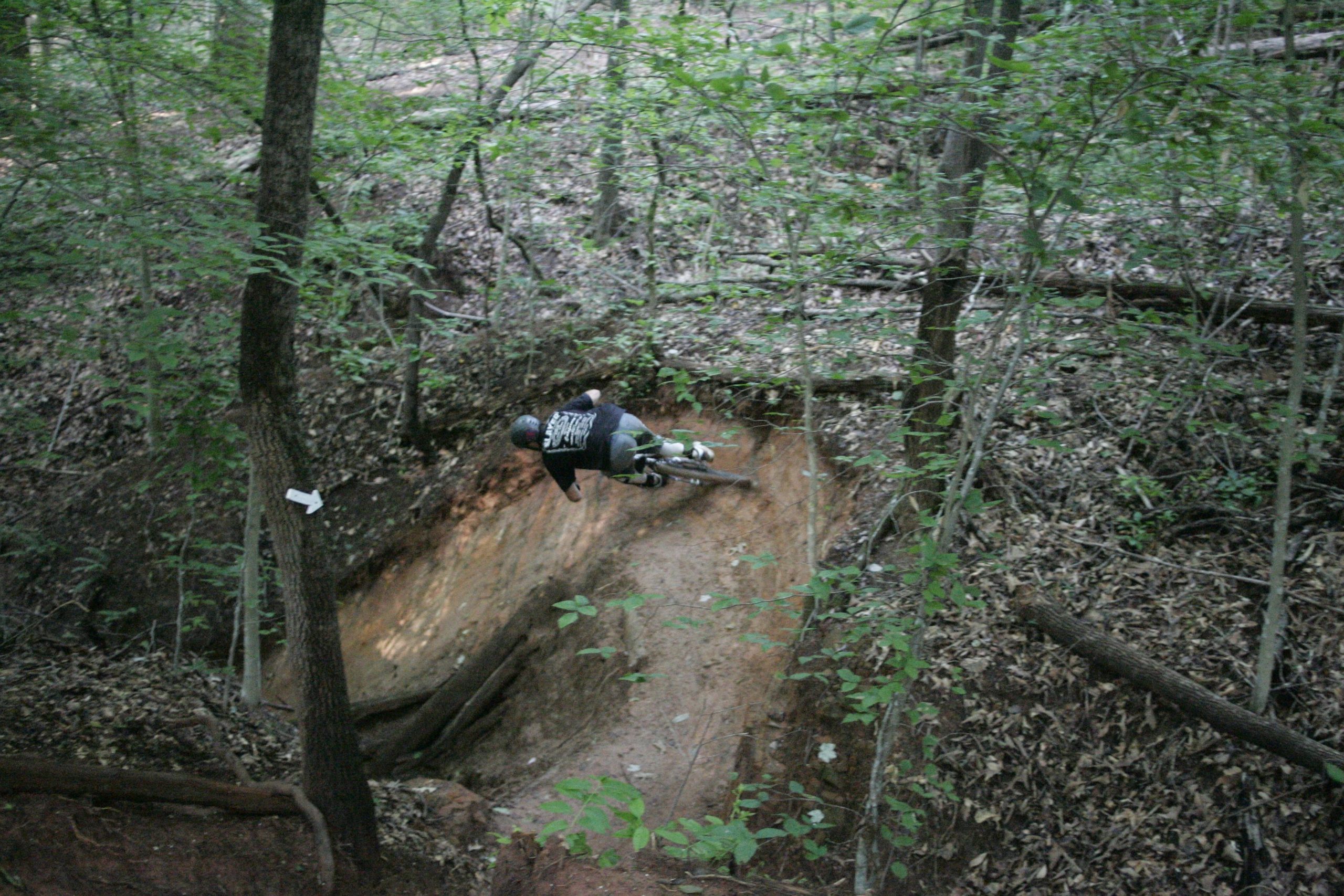 A cyclist performing a sharp turn on a dirt trail in a wooded area, surrounded by trees and foliage. The dirt path shows signs of use, and the cyclist is seen in motion, showcasing biking skills. Salem Lake mountain bike trail.