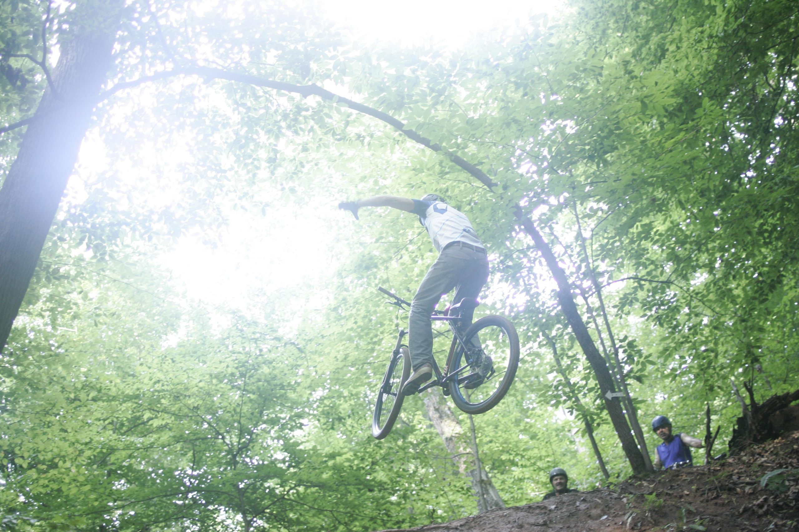 A cyclist performing a jump on a mountain bike, airborne above a dirt ramp in a green forest. Two spectators in the background watch the action. The scene is filled with lush trees and soft, diffused light. Salem Lake mountain bike trail.