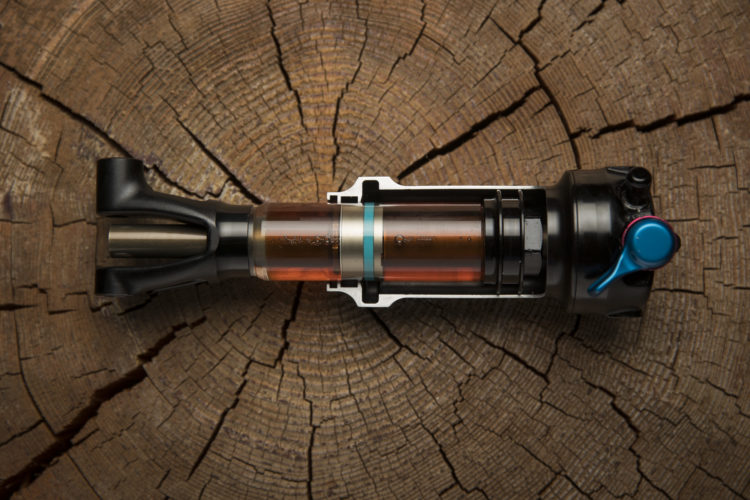 A top view of a mountain bike shock absorber displayed on a wooden surface, showing intricate details such as the outer casing, reservoir with fluid, and adjustable components. The background features a circular tree stump, highlighting natural textures.