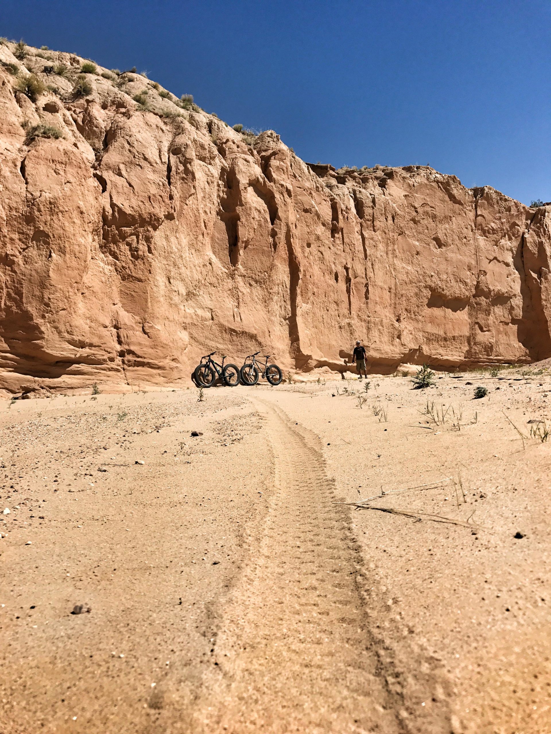 A sandy trail leads through a desert landscape, flanked by tall rock formations. In the distance, several bicycles are parked on the soft ground, with a solitary figure standing nearby under a clear blue sky. Small patches of vegetation dot the sandy terrain. Mariposa Fat Bike Trails mountain bike trail.