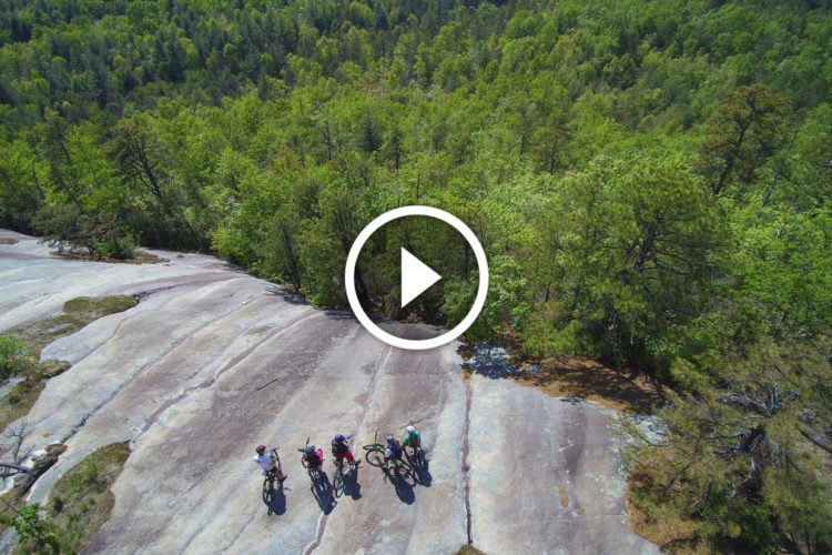 Aerial view of a group of mountain bikers standing on a rocky surface surrounded by lush green trees and vegetation. A play button overlay indicates that the image is part of a video.