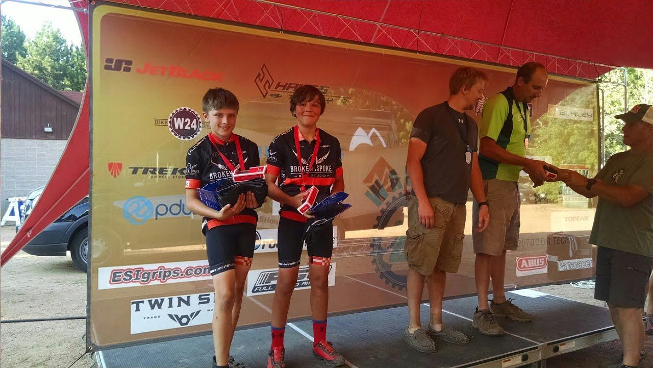 Two young cyclists wearing matching black jerseys with red accents stand on a podium, each holding blue trophies. They have gold medals around their necks and are smiling proudly. In the background, adults are handing out awards on a stage decorated with various sponsor logos. The setting appears to be an outdoor event, likely a cycling competition. Nine Mile mountain bike trail.
