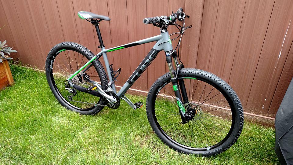 Cube ANALOG 27.5: A sleek gray mountain bike with green accents, positioned on a patch of green grass near a wooden fence. The bike features knobby tires, a sturdy frame, and a front suspension fork, showcasing its off-road capabilities.