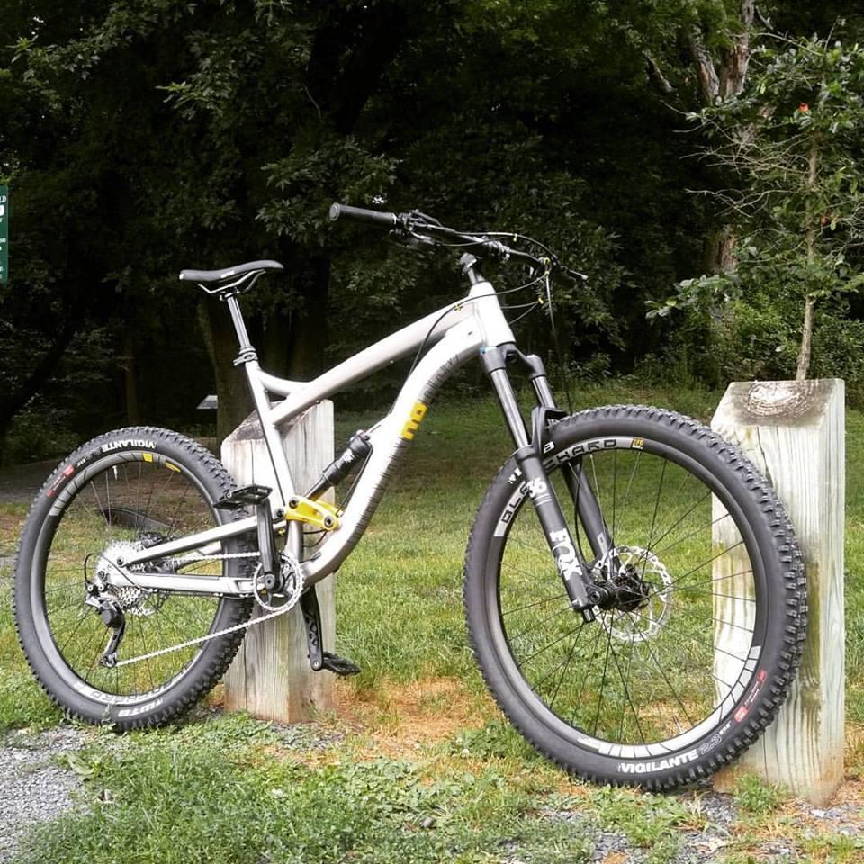 Diamondback Mission 2: A silver mountain bike leaning against a wooden post in a grassy area, surrounded by trees. The bike features thick tires, a sturdy frame, and front suspension, indicating it's designed for off-road terrain.