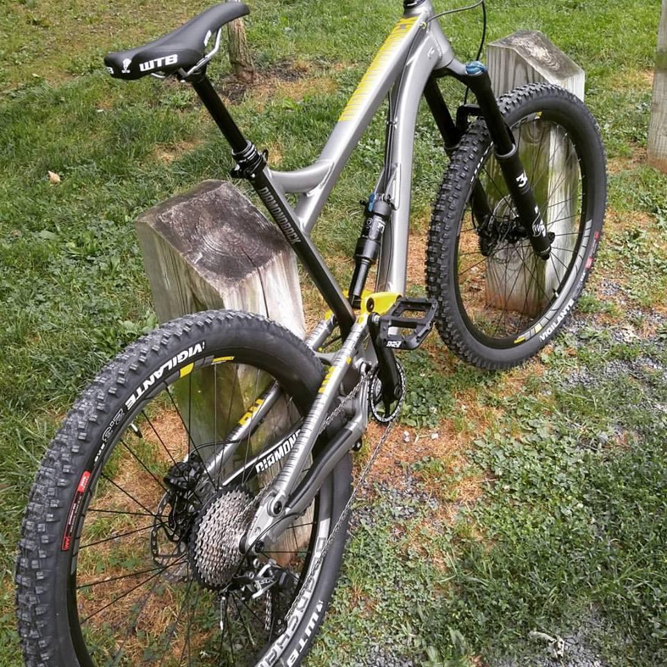 Diamondback Mission 2: A silver mountain bike with yellow accents stands next to a wooden post on a grassy area. The bike features thick tires suitable for off-road terrain, a black seat, and a suspension system designed for a smooth ride. Various components, such as pedals and gears, are visible, showcasing its advanced design for outdoor cycling.