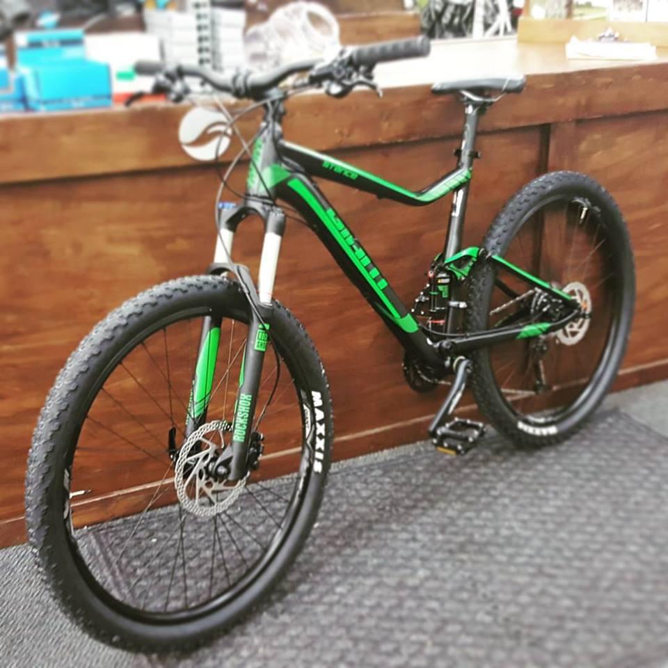 Giant Giant Stance 2: A black and green mountain bike with wide tires, positioned upright against a wooden counter. The bike features a sleek frame, suspension fork, and disc brakes, showcasing a modern design suitable for off-road cycling. The background includes various bike parts and accessories.