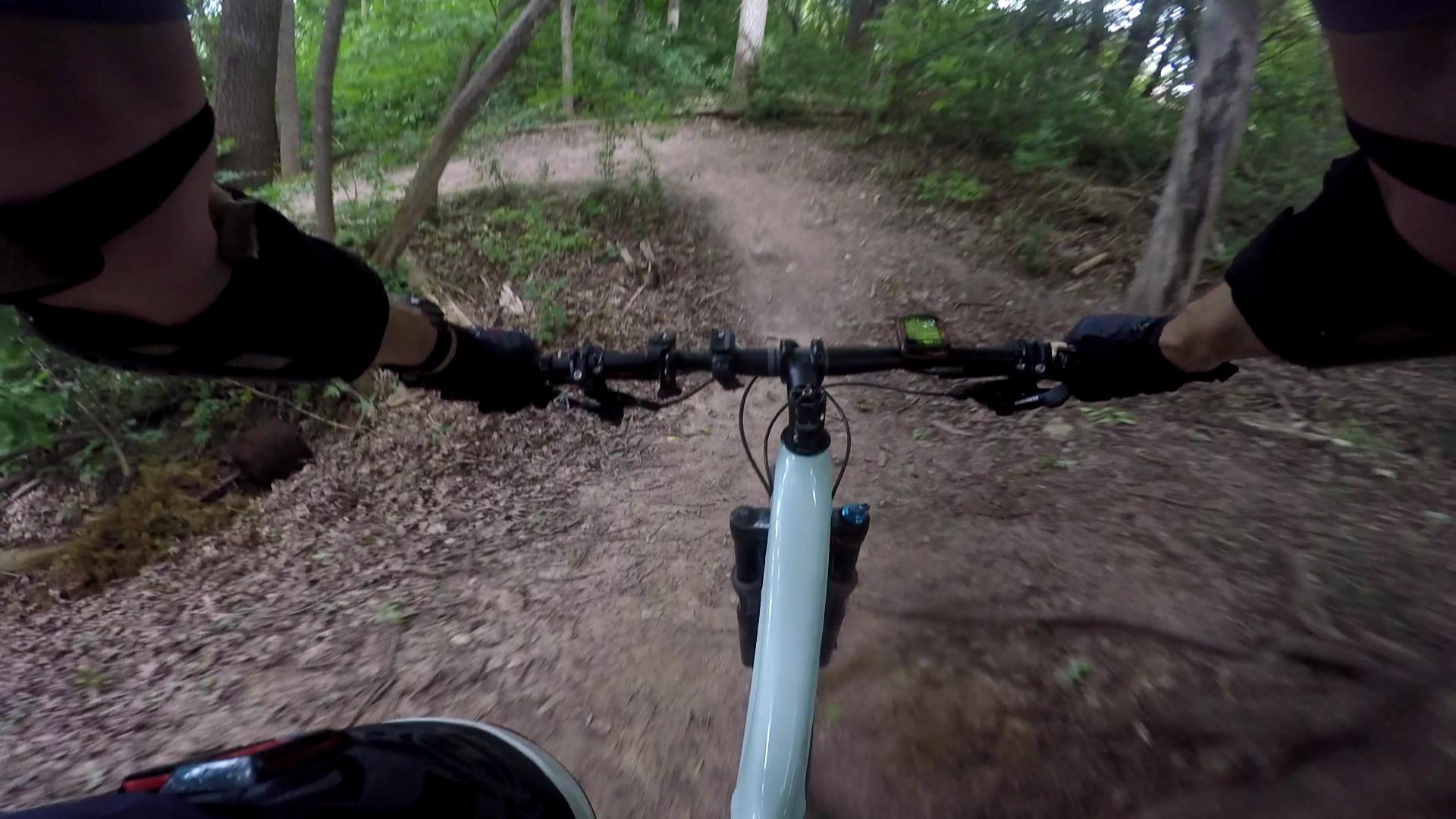 A first-person perspective of mountain biking on a dirt trail surrounded by lush greenery. The image shows the handlebars of a bike, with one hand gripping the grips and the other arm visible, wearing a glove. The trail winds ahead, featuring soft earth and scattered leaves, suggesting an active outdoor adventure. Foxtrot mountain bike trail.