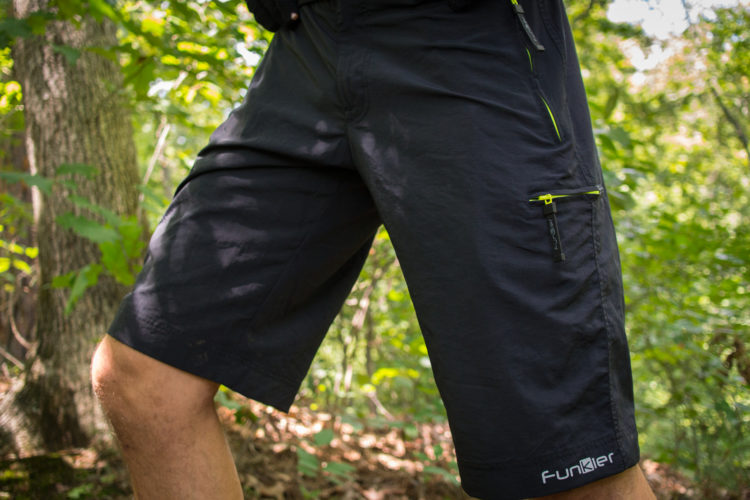Close-up of a person wearing black outdoor shorts with a zippered pocket, standing in a forested area surrounded by green foliage. The shorts feature a small logo on the hem and bright zipper accents.