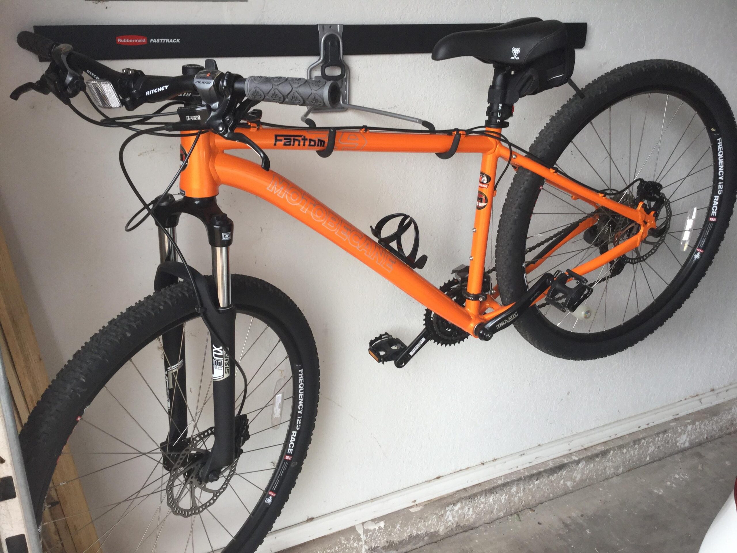 Motobecane Fantom29 Sport: A bright orange mountain bike is mounted on a wall rack in a garage. The bike features chunky tires, a front suspension fork, and a black seat. The background shows a plain wall and part of the floor, indicating a tidy storage space.