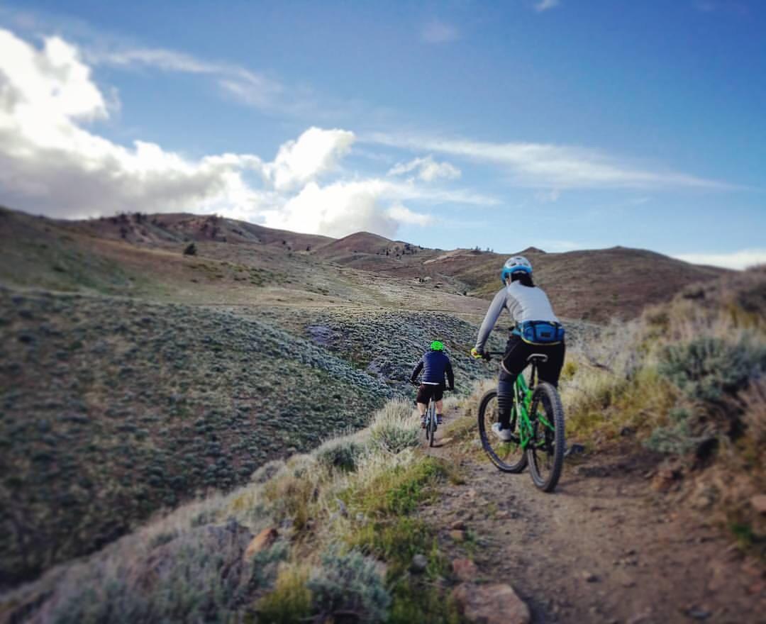 Two mountain bikers riding on a dirt trail through a grassy landscape with rolling hills under a partly cloudy sky. The rider in front is wearing a green helmet and a dark jacket, while the rider behind is in a light gray jacket and a blue helmet. Peavine mountain bike trail.