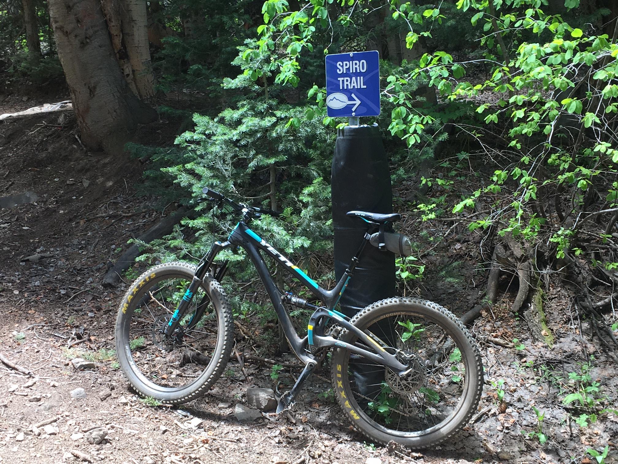 Yeti SB5c: A mountain bike resting on gravel beside a sign marking the Spiro Trail, surrounded by lush green foliage and trees.