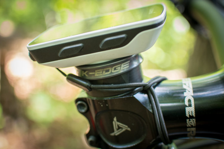 Close-up of a bike's handlebar with a K-Edge mounting system and a cycling computer attached, set against a blurred natural background.