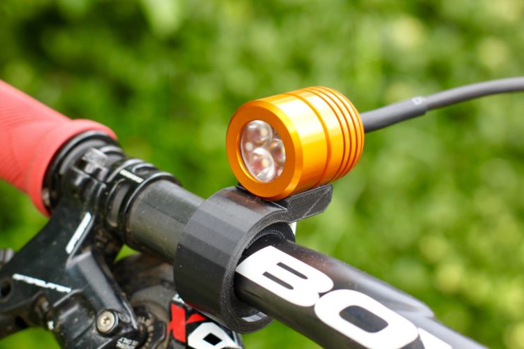 A close-up image of a bicycle handlebar featuring an orange LED light mounted securely on a black bracket. The handlebar has red rubber grips, and the background consists of greenery, suggesting an outdoor setting.