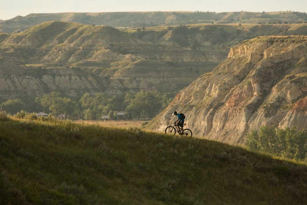 A cyclist riding a mountain bike along a grassy ridge, with rolling hills and rugged cliffs in the background under soft, natural lighting. Maah Daah Hey mountain bike trail.