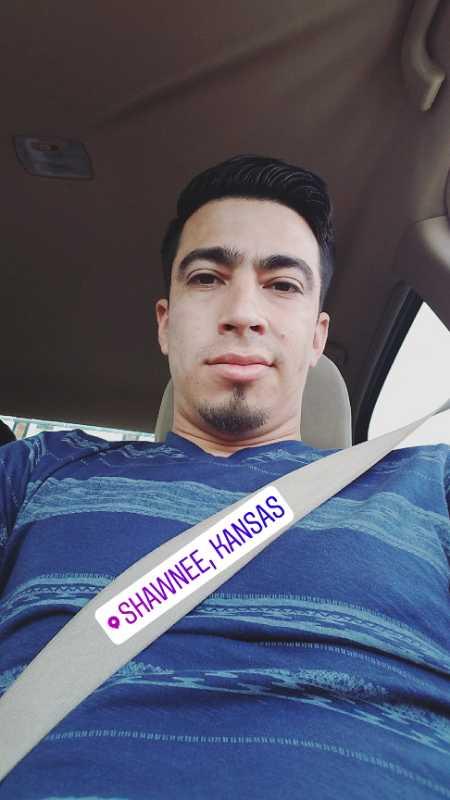 A person taking a selfie while seated in a car, wearing a blue striped shirt. A seatbelt is visible across their chest, and a location tag reading "Shawnee, Kansas" is displayed at the bottom of the image. L. F. Cosca Regional Park mountain bike trail.