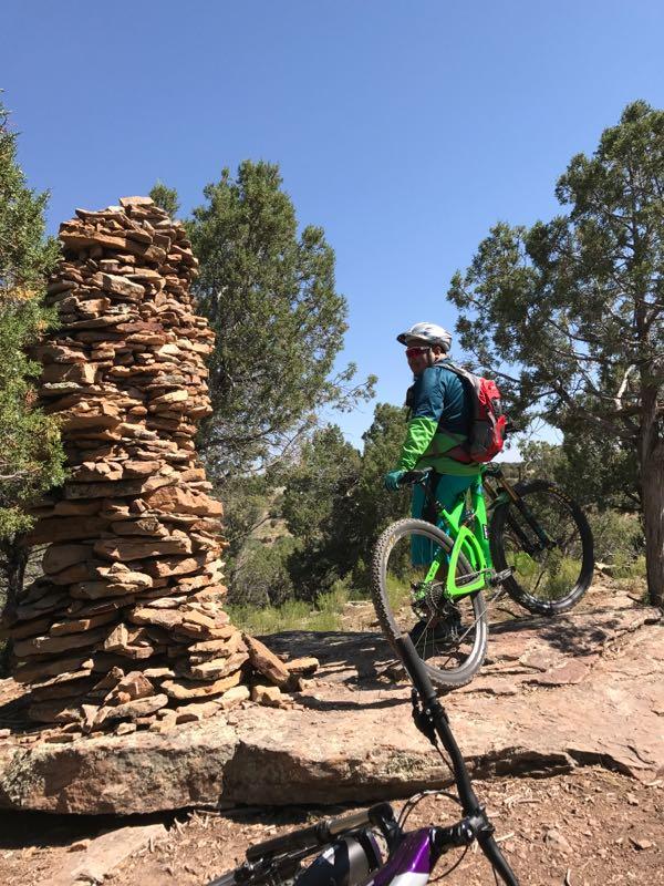 A mountain biker in a green outfit and helmet stands beside a large stone stack on a rocky trail surrounded by vegetation, with another bicycle partially visible in the foreground. The scene is set under a clear blue sky. Phil's World mountain bike trail.
