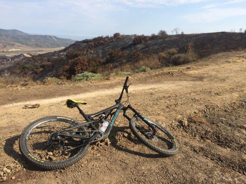 A mountain bike lying on its side on a rugged dirt path, surrounded by scorched earth and sparse vegetation. In the background, a hilly landscape is visible under a clear blue sky. San Clemente Singletracks mountain bike trail.