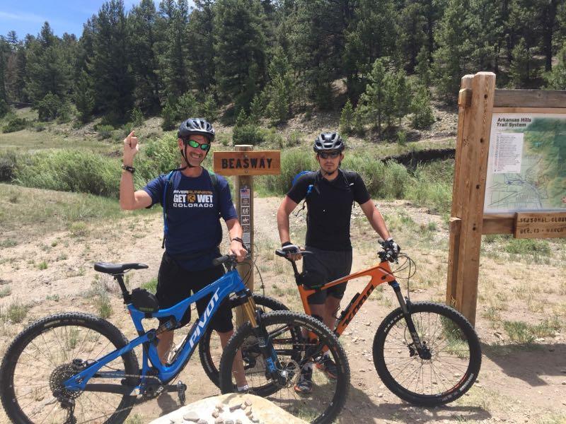 Two mountain bikers stand next to their bikes at a trailhead marked "Beasway." One rider, wearing a helmet and sunglasses, raises a hand in excitement, while the other stands beside him, both smiling. The background features green trees and a trail map of the Arkansas Hills Trail System. Cottonwood mountain bike trail.