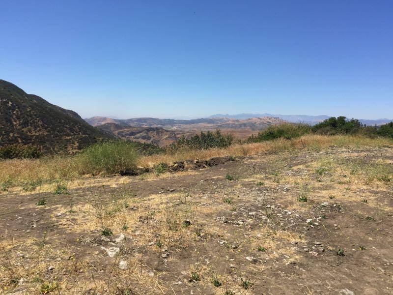 A panoramic view of a hilly landscape under a clear blue sky, featuring dry grass, patches of greenery, and distant mountains in the background. The foreground shows a rocky terrain with sparse vegetation. Las Llajas Canyon mountain bike trail.