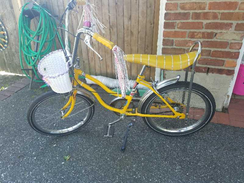 A vintage yellow bicycle with a unique design, featuring a curved frame, a striped seat, and a white wicker basket attached to the handlebars. The bike is parked on a paved surface near a wooden fence and garden hose. Meadowlark Park mountain bike trail.