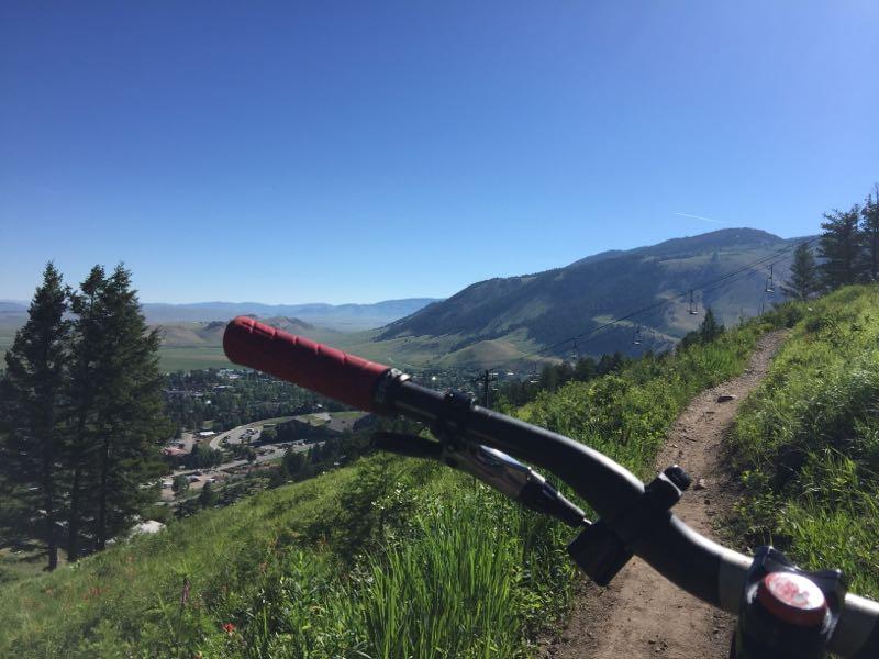A view from a mountain bike on a scenic trail, overlooking a valley and town below, surrounded by green hills and trees under a clear blue sky. Snow King Mountain mountain bike trail.