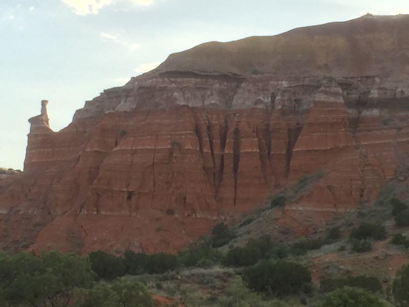 A close-up view of a dramatic rock formation, showcasing layers of red and orange sandstone cliffs. The cliffs are textured with vertical grooves and ledges, surrounded by sparse green vegetation at the base. The sky above is slightly cloudy, giving a scenic backdrop to the geological features. Palo Duro Canyon mountain bike trail.
