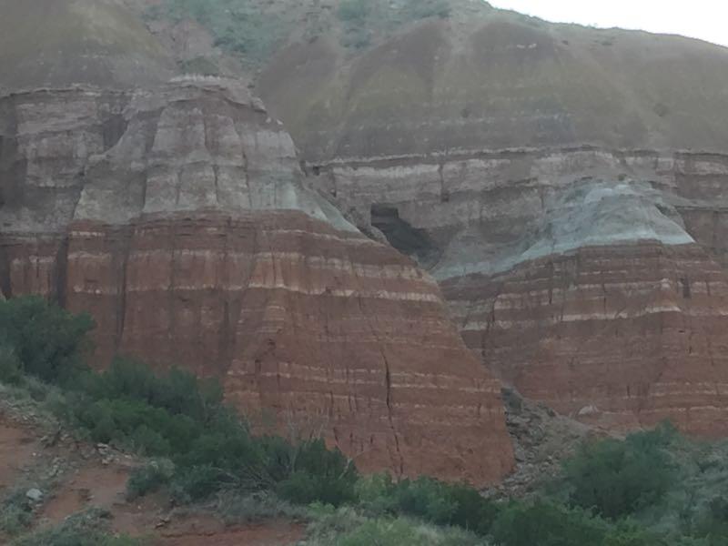 Alt text: A landscape featuring layered rock formations in various shades of red, brown, and gray, with vegetation at the base, set against a backdrop of hills. Palo Duro Canyon mountain bike trail.