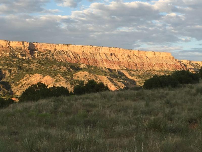 A scenic view of colorful rock formations rising sharply against a backdrop of clouds, with lush green grass and bushes in the foreground. The layered textures of the cliffs are highlighted by the warm sunlight, showcasing shades of orange, red, and beige. Palo Duro Canyon mountain bike trail.