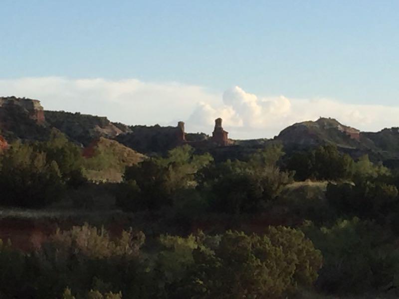 A scenic view of rugged rock formations in a landscape filled with sparse vegetation, under a partly cloudy blue sky. The image captures the natural beauty of the terrain, with distant cliffs and a serene atmosphere. Palo Duro Canyon mountain bike trail.