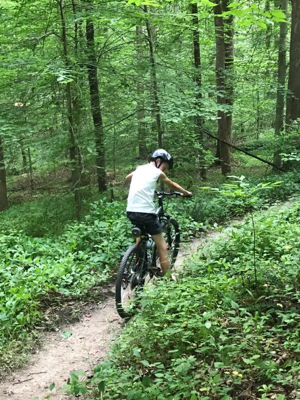 A child riding a mountain bike along a narrow dirt trail through a lush, green forest, surrounded by trees and underbrush. Rosaryville State Park mountain bike trail.