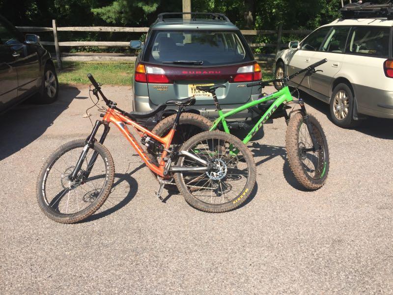 Two mountain bikes, one orange and one green, are leaning against each other in a parking area. In the background, a Subaru vehicle is parked, along with another car. The ground is paved with gravel, and trees are visible in the distance, suggesting an outdoor setting. Six Mile Run mountain bike trail.