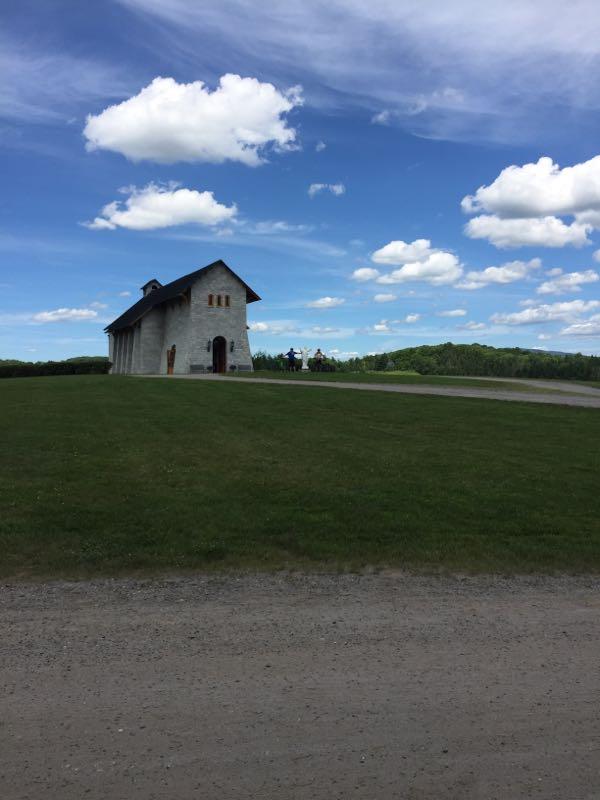 A scenic view of a rustic building on a grassy hill, with a clear blue sky overhead featuring white clouds. The building has a sloped roof and is constructed from stone, with a few figures visible in front of it. In the background, rolling green hills can be seen. Kingdom Trails mountain bike trail.