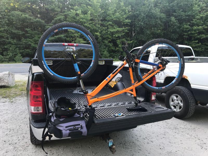 A mountain bike with an orange frame and blue wheels is secured in the bed of a pickup truck, with one wheel resting upright. A black helmet and a small purple bag are positioned beside the bike. In the background, trees and another vehicle are visible. Range Pond State Park Trails mountain bike trail.