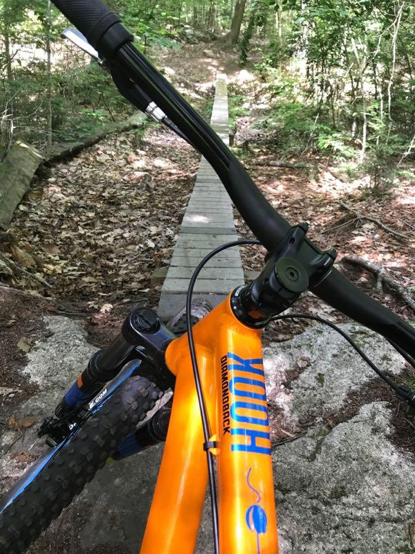 A close-up view of a mountain bike's handlebars and frame, featuring a bright orange color, positioned on a narrow wooden bridge trail surrounded by a forest with fallen leaves. The perspective is from the bike's point of view, looking down the path ahead. Bradbury Mt State Park mountain bike trail.