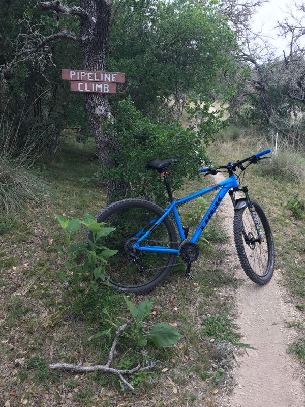A blue mountain bike is leaned against a tree with a wooden sign that reads "Pipeline Climb." The scene is surrounded by greenery and a dirt trail. Flat Rock Ranch mountain bike trail.