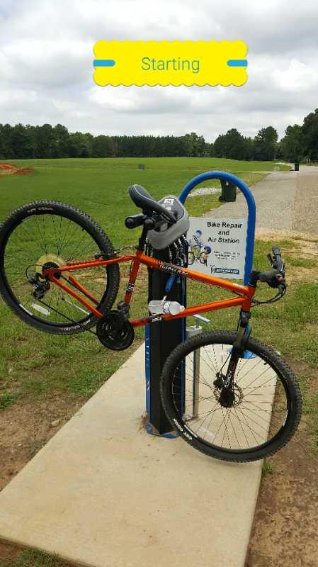 An orange bicycle is mounted on a bike repair and air station, set against a grassy field and cloudy sky. The station features tools for bike maintenance and an air pump. A sign above indicates the starting point for cyclists. Beaver Creek / Forever Wild Trails mountain bike trail.