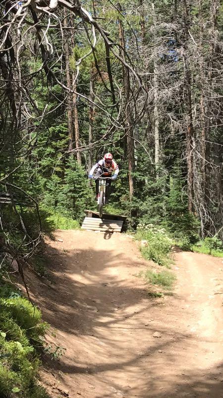 A mountain biker performing a jump off a wooden ramp, surrounded by lush green trees and a dirt trail, with sunlight streaming through the forest. Trestle Bike Park mountain bike trail.