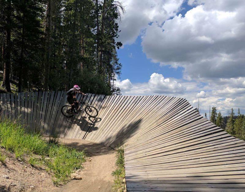 A mountain biker navigates a wooden pump track while surrounded by evergreen trees under a partly cloudy sky. The track features a wavy design, allowing for dynamic movement and jumps as the cyclist rides along the curvy surface. Trestle Bike Park mountain bike trail.