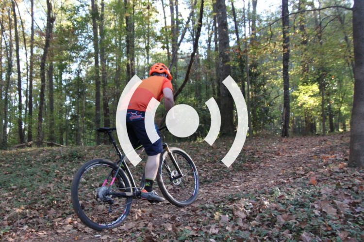 A cyclist in bright orange gear rides a mountain bike along a dirt trail surrounded by trees, with autumn leaves scattered on the ground. The image features a design overlay indicating sound waves.