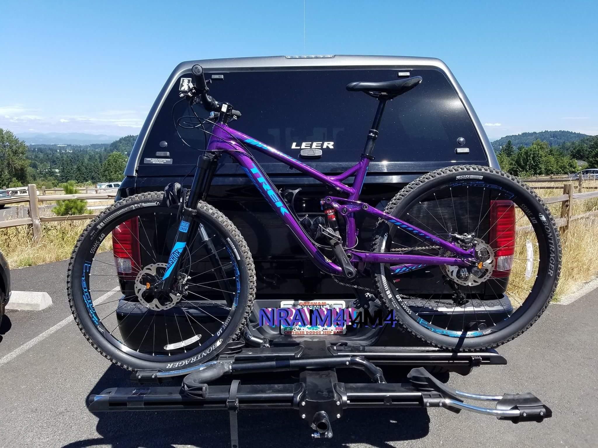 Trek Remedy 9 27.5/650b: A purple mountain bike secured to a bike rack on the back of a vehicle, parked in a scenic area with green hills and a blue sky in the background. The bike features prominent branding and has knobby tires designed for off-road riding.
