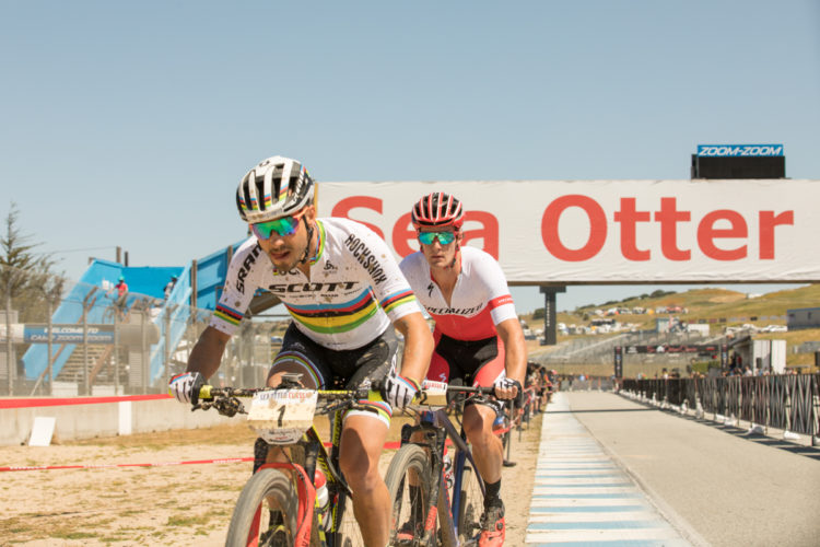 Two competitive cyclists race towards a large banner reading "Sea Otter" at an outdoor event. The cyclist in the front wears a rainbow-striped jersey and sunglasses, while the cyclist behind him is in a red and white jersey. The background features a clear blue sky and spectators lining the course, with the race track visible.