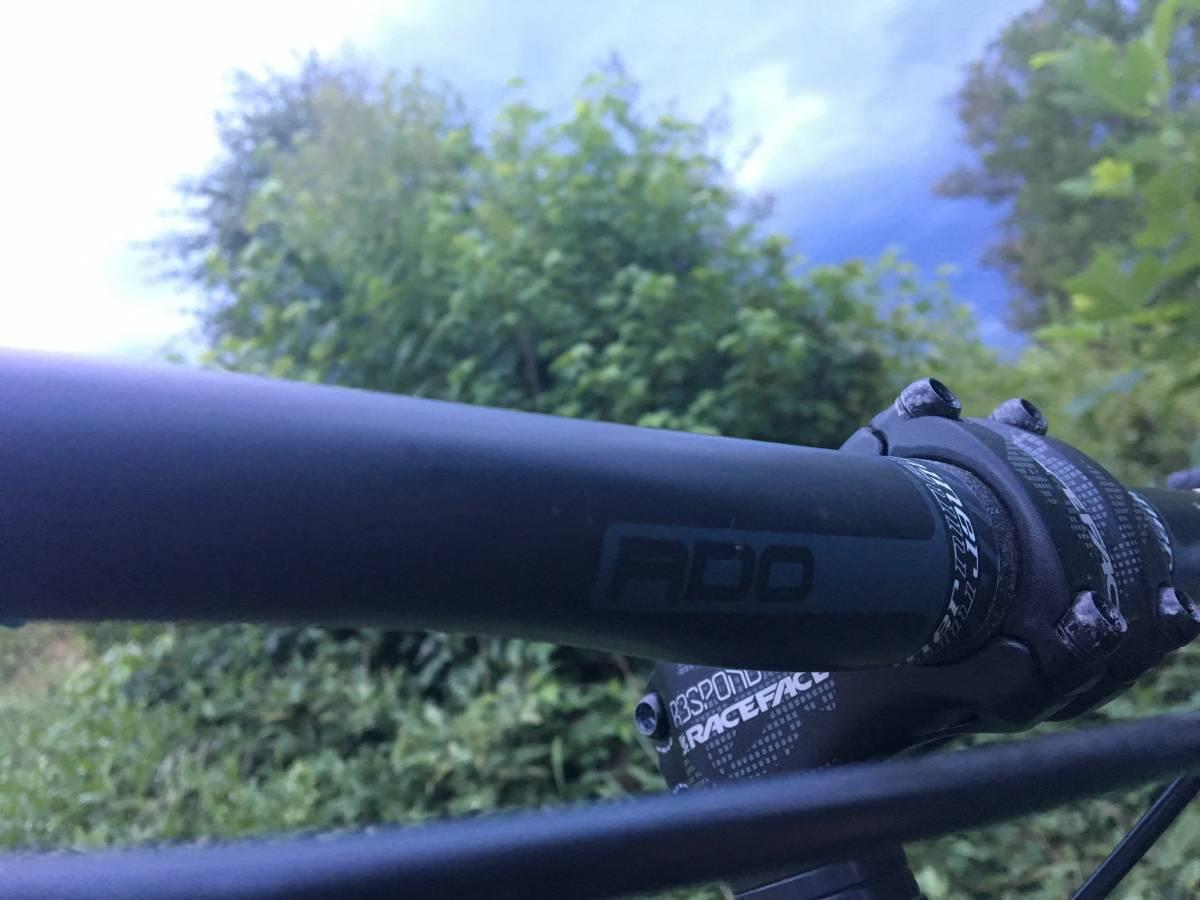 Kona Process 111: Close-up of a bicycle handlebar showing the ADO branding, with a blurred background of lush green foliage and a cloudy sky.