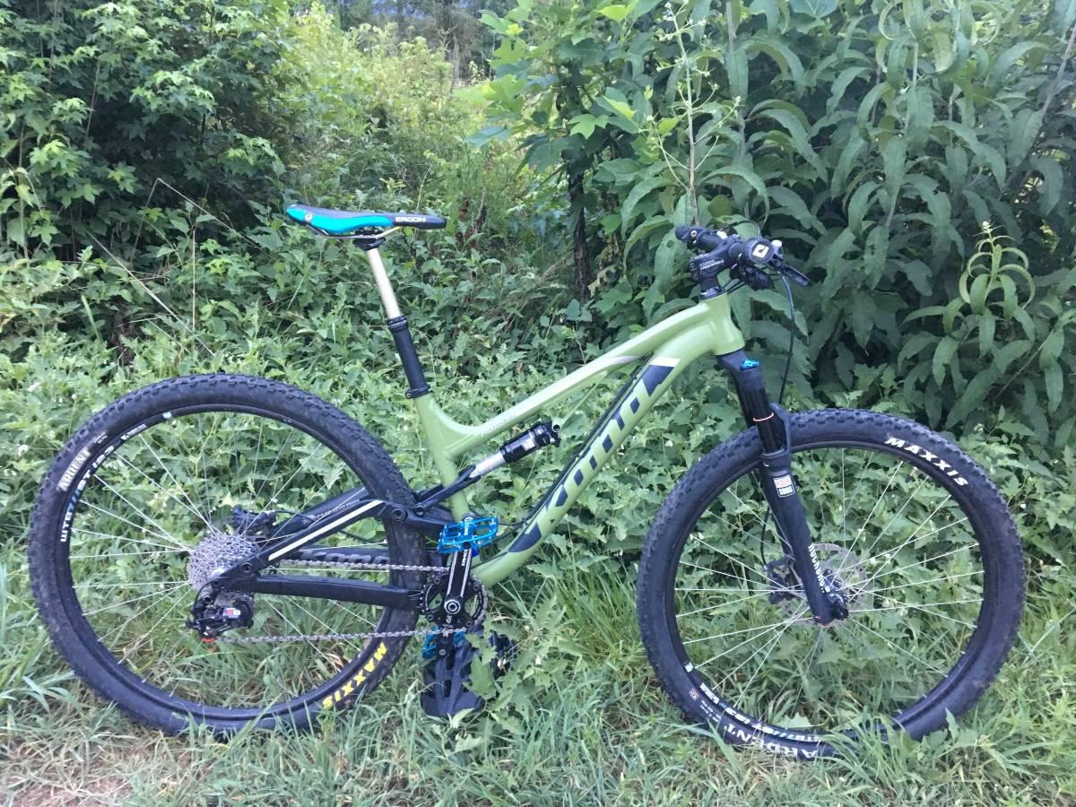 Kona Process 111: A green and black mountain bike rests on grass near dense foliage, showcasing its sturdy frame, large tires, and high-performance components. The bike's wheels are equipped with Maxxis tires, and it features a blue and black saddle. The setting suggests a natural outdoor environment, ideal for biking adventures.