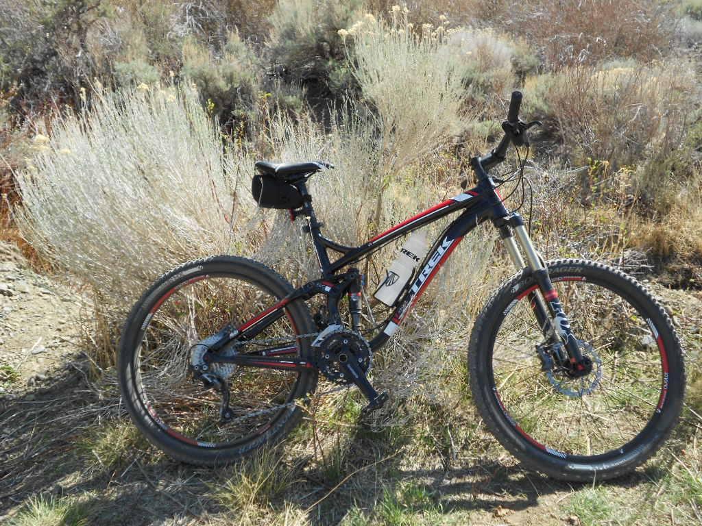 Trek Fuel EX 7: A mountain bike leaning against a bush in a natural outdoor setting, with dry grass and shrubs in the background. The bike is primarily black with red accents and features a rear bag attached to the seat.