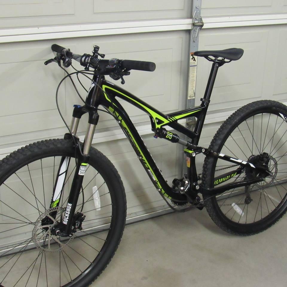 Specialized Camber 29: A black and green mountain bike is leaning against a garage wall. The bike features a sleek frame, front suspension, and knobby tires suitable for off-road cycling. The handlebars are equipped with grips and controls, and the bike has disc brakes visible on the wheels.