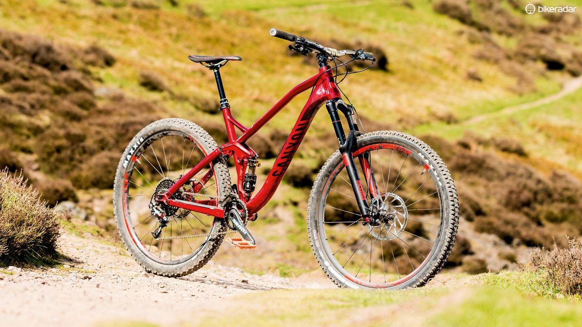 Canyon Strive CF: A red Canyon mountain bike positioned on a gravel path surrounded by green hills and heather. The bike features thick tires and a sturdy frame, designed for off-road cycling.