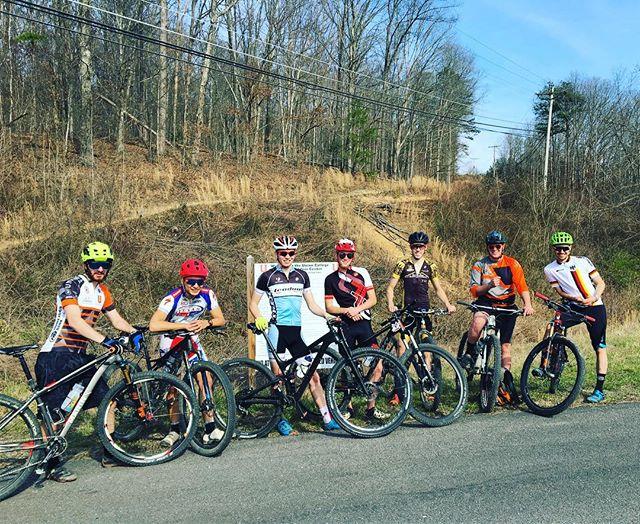 A group of seven cyclists posing for a photo beside their mountain bikes on a sunny day. They are dressed in various cycling jerseys and helmets, standing on a rural road with dry grass and trees in the background. Turner Outdoor Center mountain bike trail.