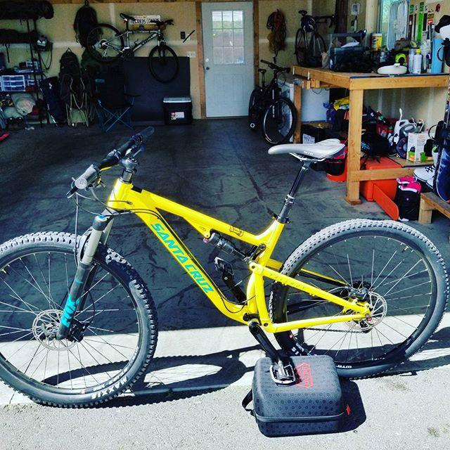 Santa Cruz Tallboy: A bright yellow mountain bike with the brand name "Santa Cruz" visible on the frame, positioned in a garage. The background features various biking accessories, tools, and other bicycles hanging on the wall. Sunlight filters into the space, highlighting the bike and its surroundings.